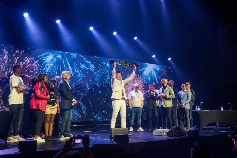 Un grupo de personas en el escenario sostiene un trofeo, celebrando su victoria con sonrisas y aplausos del público.